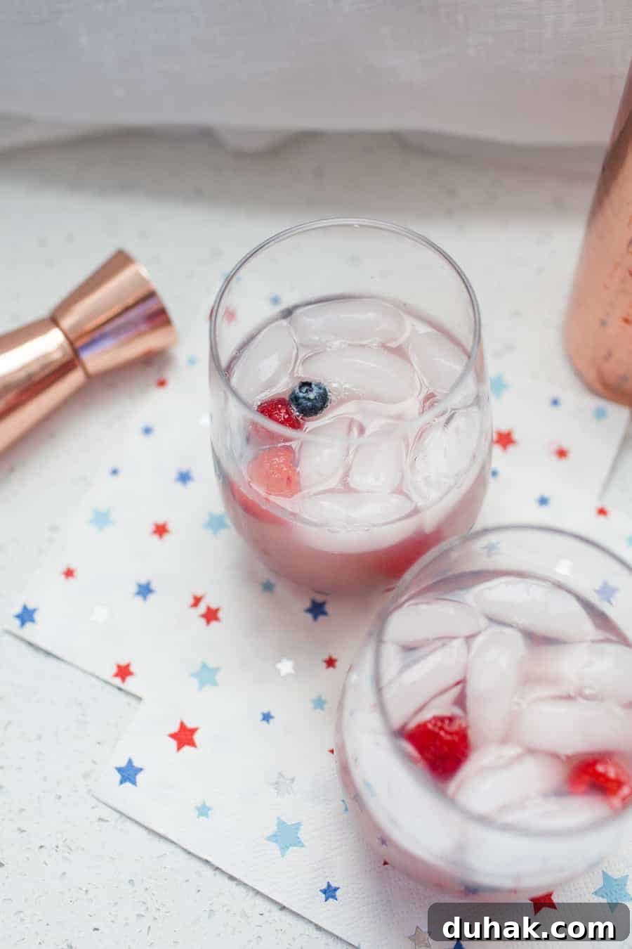 Classic Tequila Soda Recipe: Crisp Low-Calorie Cocktail Guide 8 Two glasses filled with ice, clear liquid, and berries sit on a napkin with star patterns. A jigger and a part of a copper-colored container are adjacent to the glasses, ready to concoct a refreshing White Wine Spritzer.