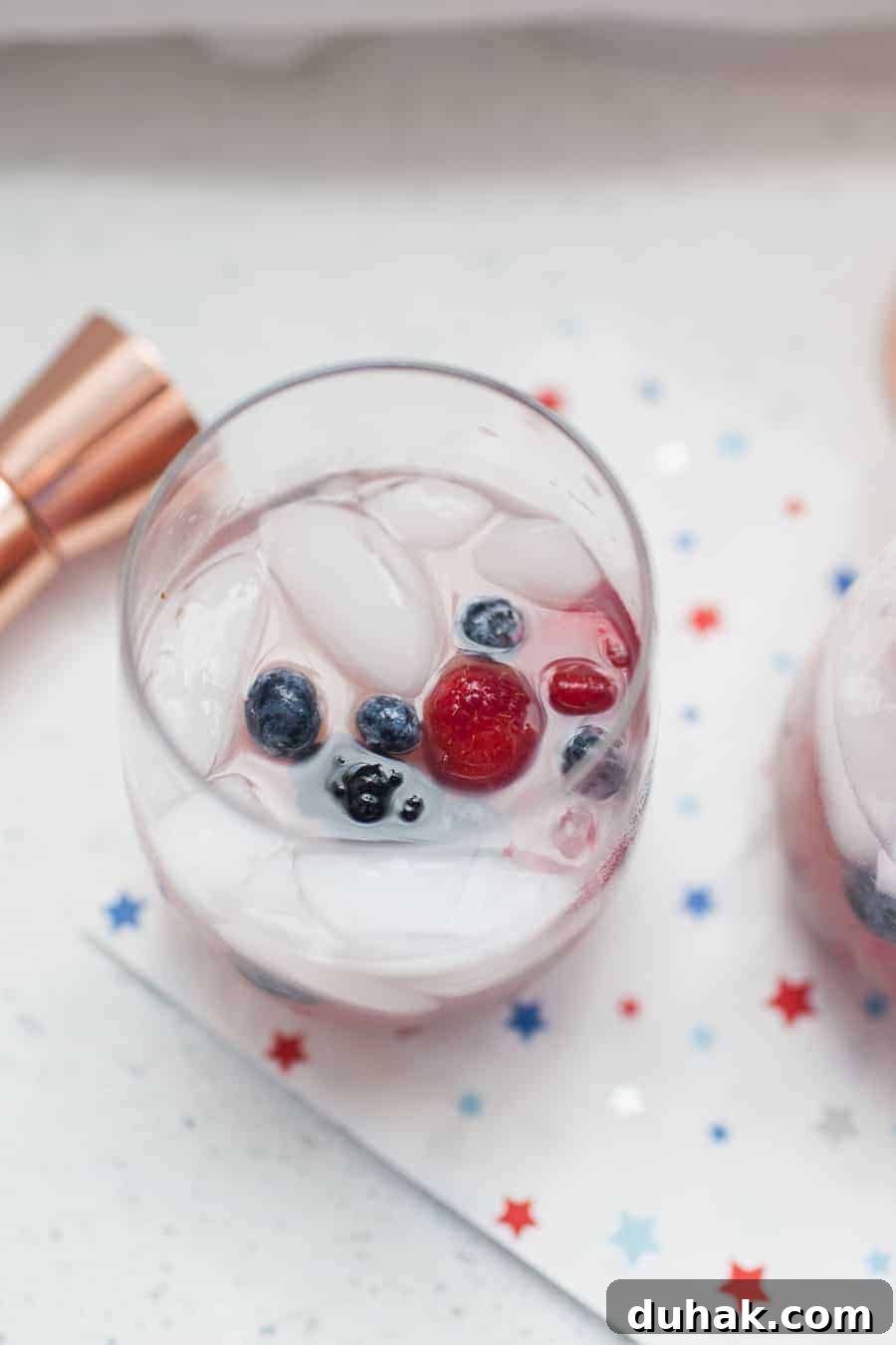 A chilled glass of White Wine Spritzer garnished with blueberries and a raspberry, placed on a white napkin with blue and red stars.